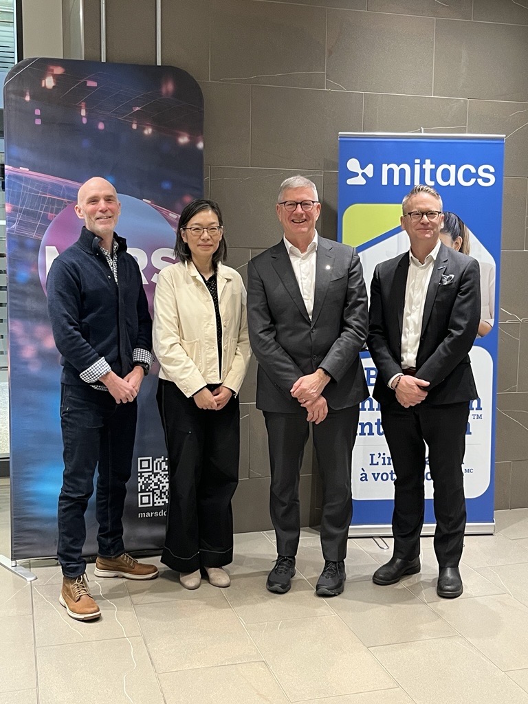 Four people stand side by side indoors, smiling at the camera. Behind them are two vertical banners, one with the Mitacs logo and another with graphics and text. The setting appears formal or professional.