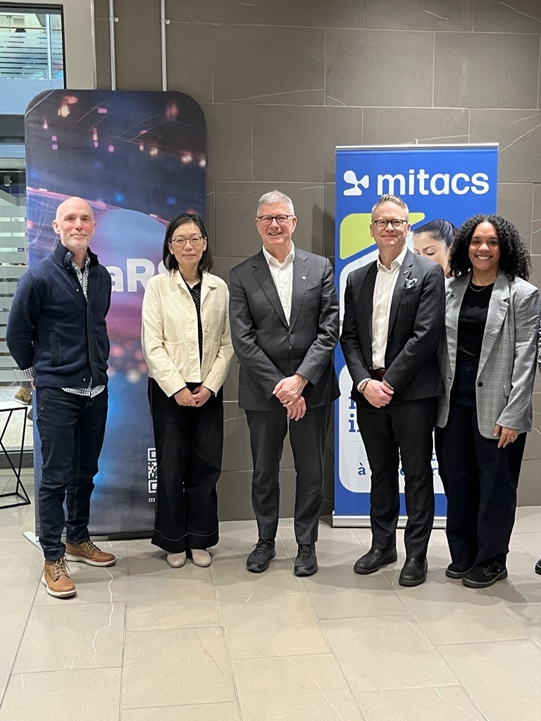 Five people stand side by side, smiling in business attire, in front of two banners—one with a colorful abstract design and the other with a blue mitacs logo and text. They are indoors on a tiled floor.
