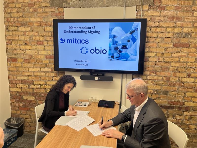 Two people sit at a table signing documents. Behind them, a screen displays “Memorandum of Understanding Signing, Mitacs Southeast College partner for research collaboration, December 2023, Toronto, ON.” The office features a brick wall backdrop.