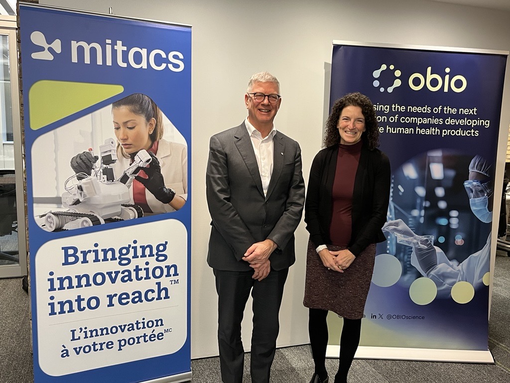 Two people stand smiling between banners for Mitacs and Obio, celebrating the new Mitacs Southeast College partner for research collaboration. The Mitacs banner shows a woman with robotics; the Obio banner highlights support for human health product companies.