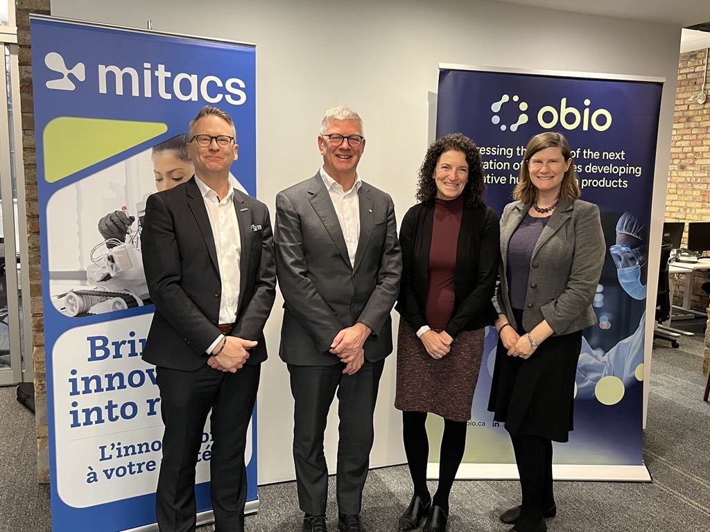 Four people in business attire stand and smile in front of banners for Mitacs and Obio, highlighting the Mitacs Southeast College partner for research collaboration, in a modern office with a brick wall and computer in the background.