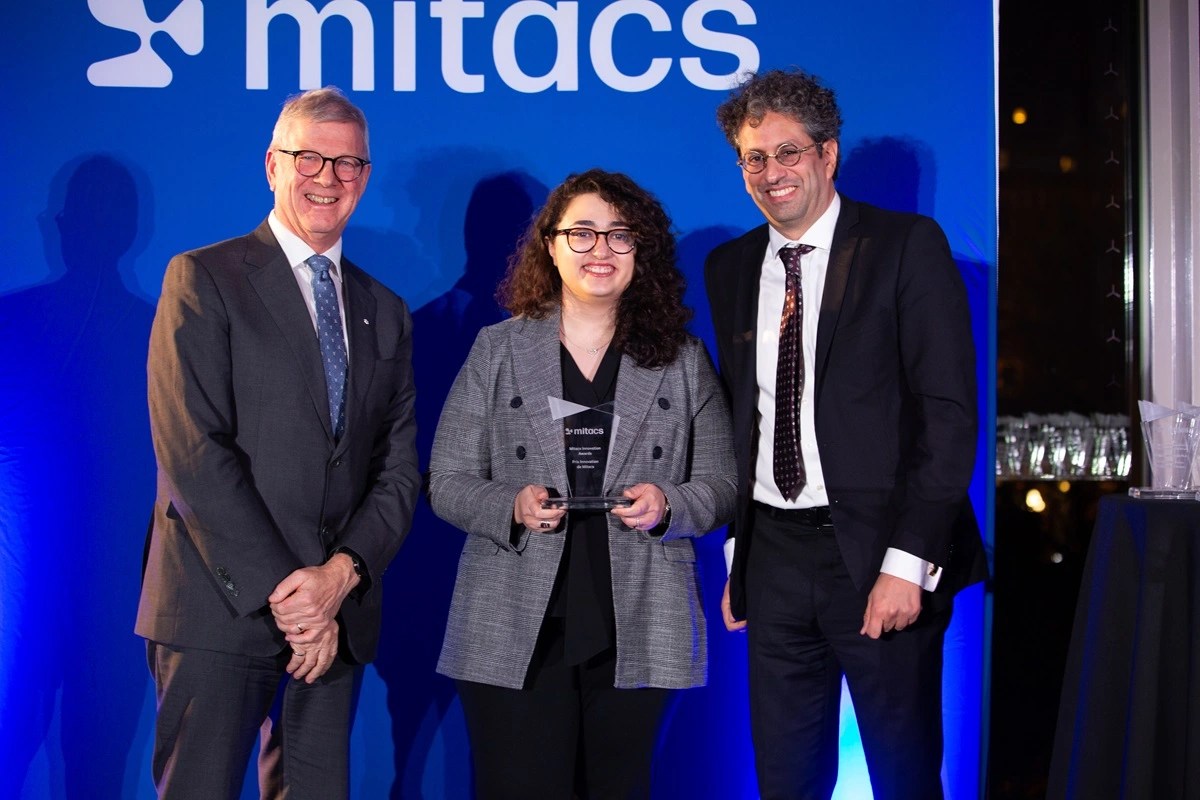 Three people in business attire stand smiling in front of a blue background with the word Mitacs; the person in the center holds an award.
