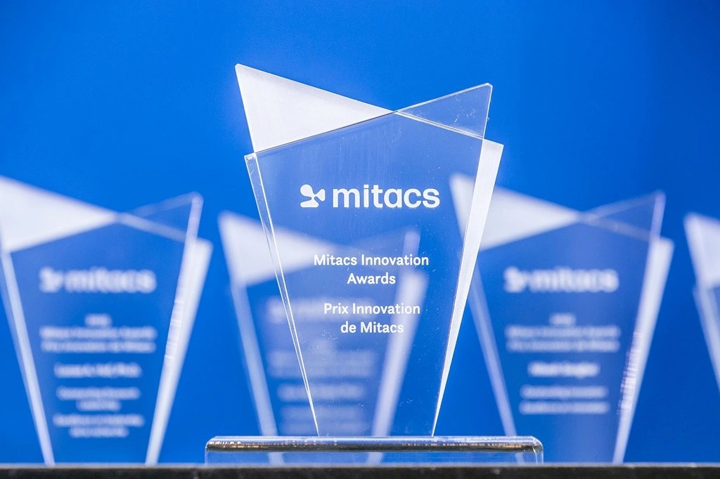 A clear glass award engraved with Mitacs Innovation Awards and Prix Innovation de Mitacs stands in front of a blue background, with several similar awards visible in the background.