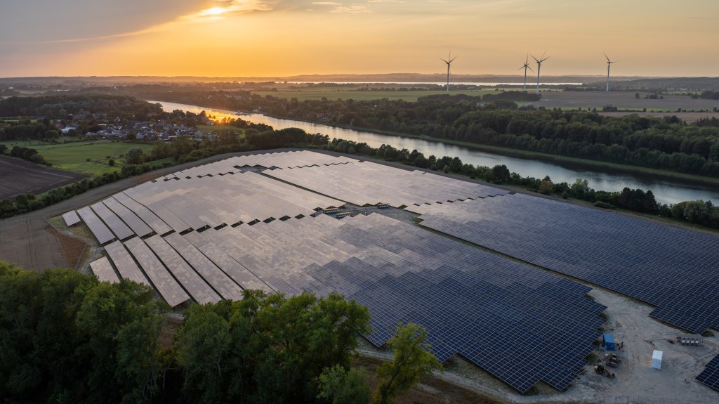 A large solar farm with rows of solar panels is situated near a river at sunset, surrounded by trees and fields, with wind turbines and a distant village visible in the background.
