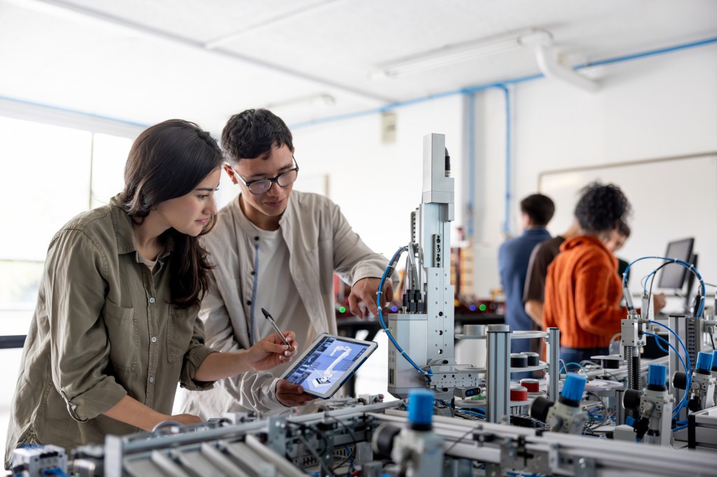 Two young adults work together on a tablet and a robotic machine in a technical lab, while two others collaborate in the background among various pieces of equipment.