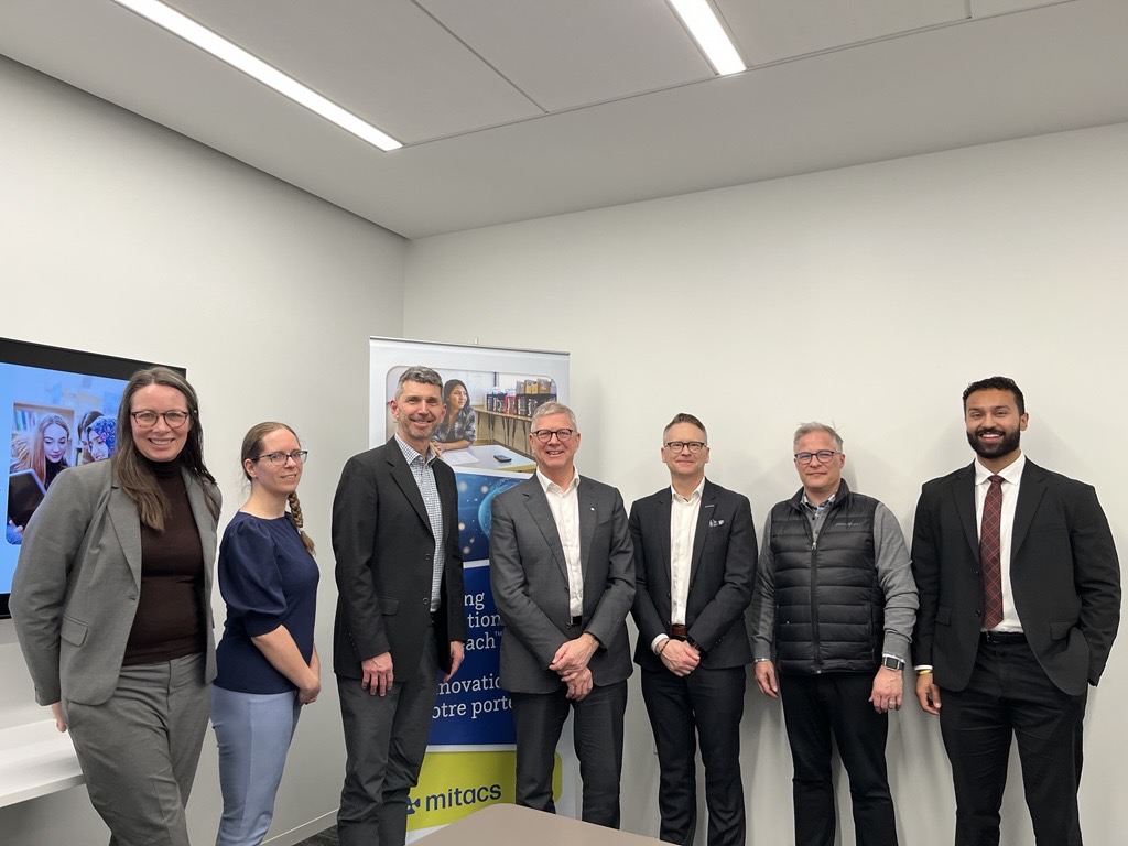 Seven people, dressed in business attire, stand side by side in a conference room. Behind them is a banner with the Mitacs logo and a screen displaying images, highlighting the Mitacs Strategic Plan 2026-2030: Talent and Research Powering Innovation.