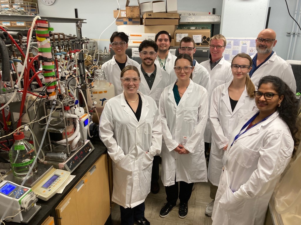 A group of ten people wearing white lab coats and safety glasses stand and smile together in a laboratory filled with scientific equipment, wires, and bottles.