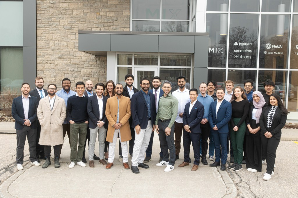 A group of 26 people, dressed in business casual and formal attire, stand together smiling in front of a modern building entrance with glass doors and signage.