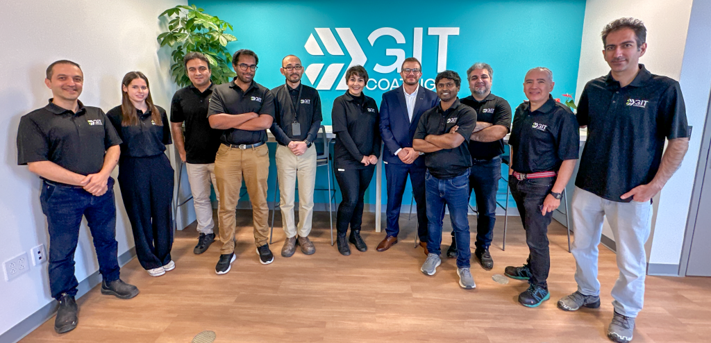 A group of twelve people stands smiling in an office with wooden floors, all wearing black shirts with 3IT logos, in front of a blue wall with the 3IT logo and the word COACHING.