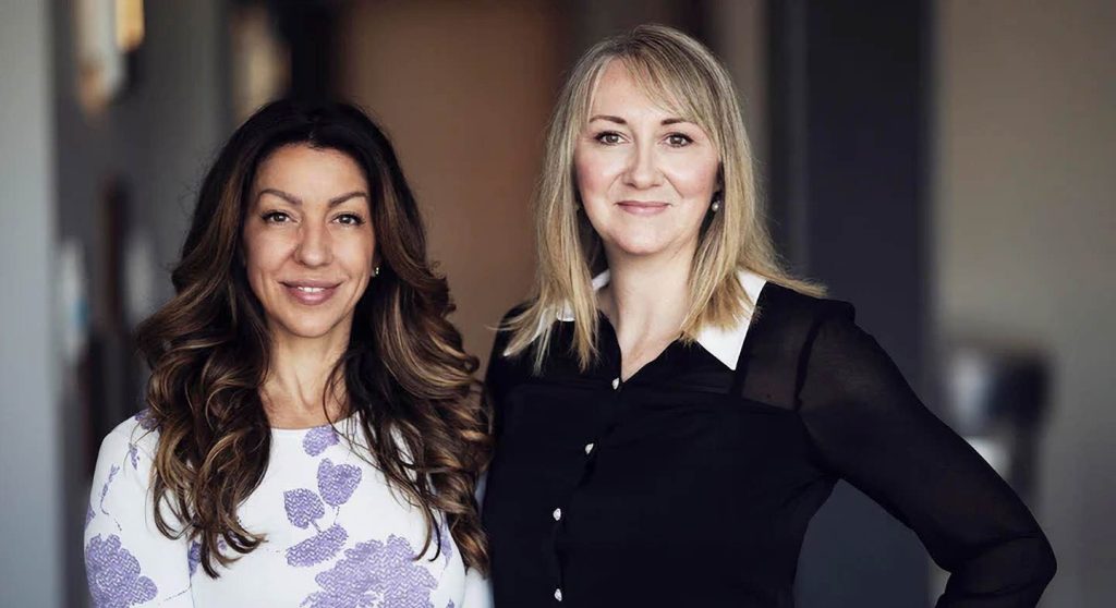 Two women standing side by side indoors, smiling at the camera. One wears a white dress with purple floral patterns; the other wears a black blouse with white accents. The background is softly blurred.