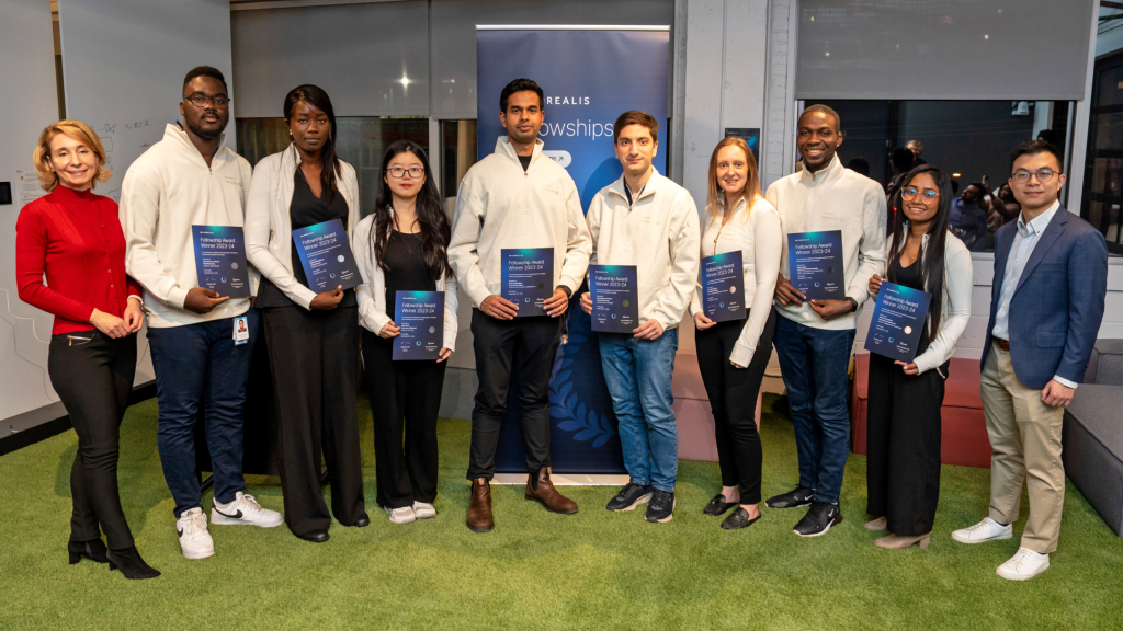 A group of ten people stand indoors in a line, some holding certificates and smiling at the camera. They appear to be participants in a fellowship program, with a banner behind them that reads Fellowships.