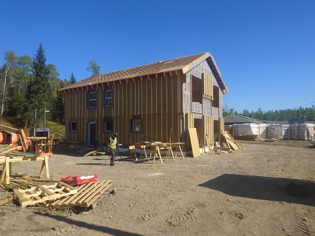 A partially constructed two-story wooden house stands on a dirt lot, surrounded by construction materials, wooden pallets, and work tables. A person in a safety vest is walking nearby under a clear blue sky.