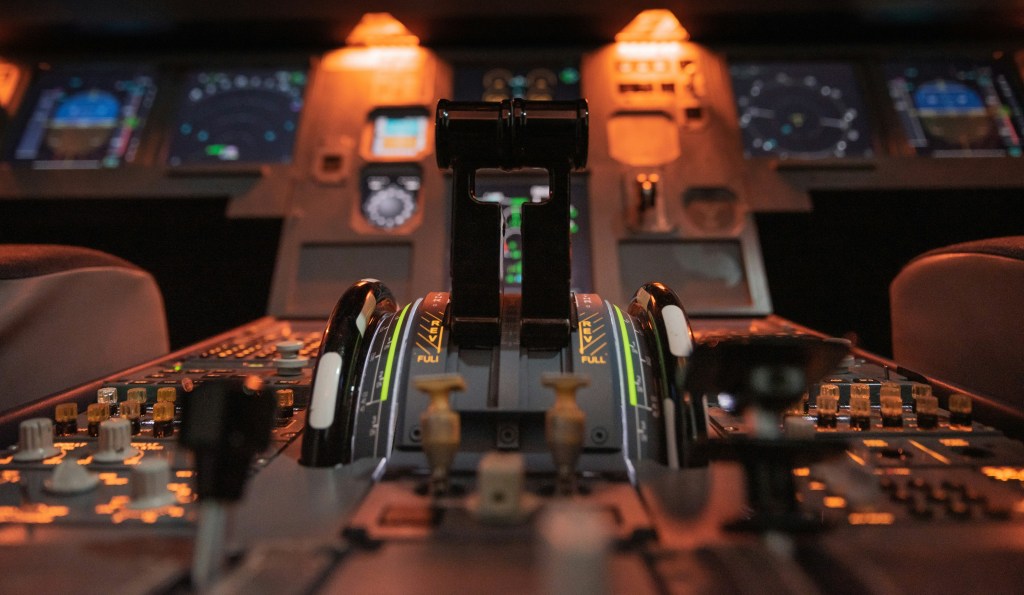 Close-up view of an illuminated airplane cockpit, focusing on the throttle controls and instrument panels, with multiple screens and buttons visible, bathed in orange and blue lights.