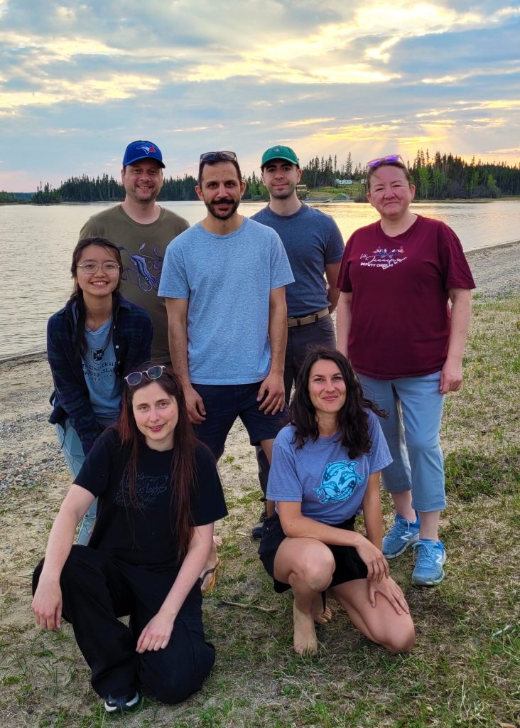 Seven people pose and smile on a lakeshore at sunset, with trees and calm water in the background—gathered as part of efforts by Cree coastal communities to improve food security and protect fish along these peaceful shores.
